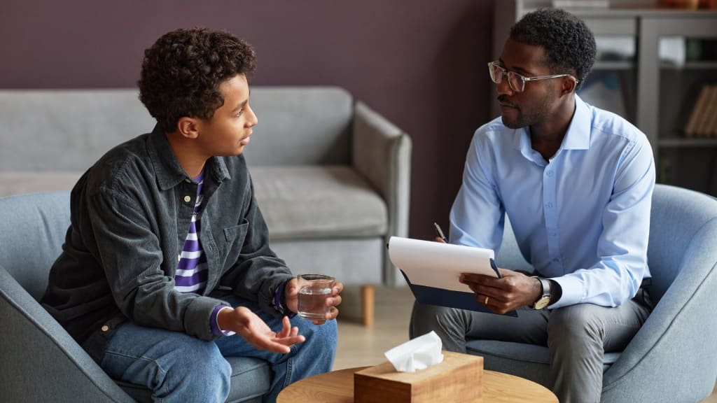 A man in glasses holds a clipboard, conversing with a seated teenager holding a glass. They are in a room with gray furniture and a tissue box on the table between them.